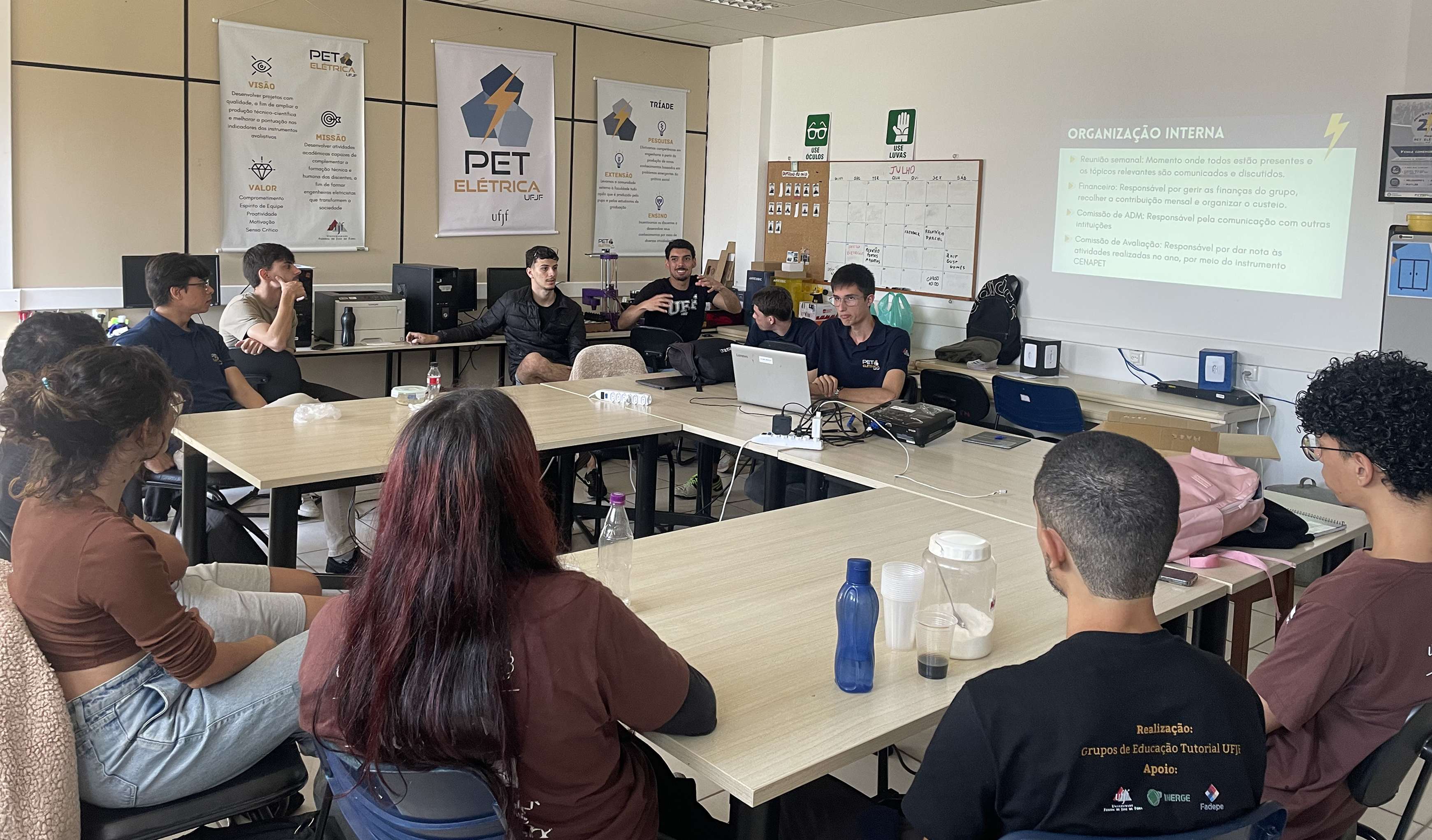 A foto mostra 11 pessoas reunidas em volta de uma grande mesa. Ao fundo, é possível observar a transmissão de um slide e também de banners do PET Elétrica.